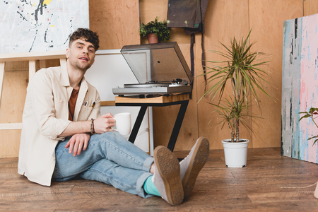 handsome artist holding coffee cup and looking at camera while sitting near vinyl record  playerの写真素材