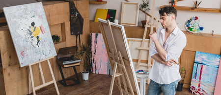 panoramic shot of handsome artist standing at easel in painting studioの写真素材