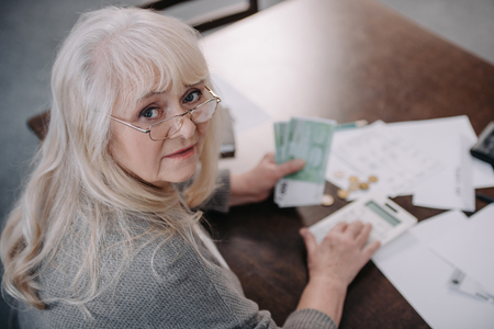 senior woman sitting at table, looking at camera, using calculator and counting moneyの写真素材