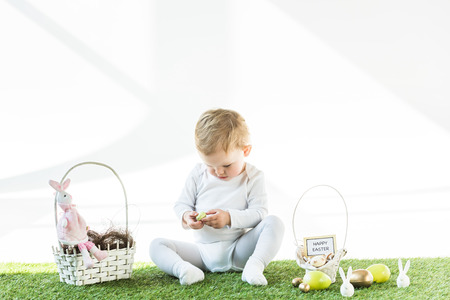 cute kid sitting near straw baskets with Easter eggs, bunnies and happy Easter card isolated on...