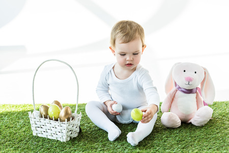 adorable child holding colorful chicken eggs while sitting near straw basket and toy rabbit isolated...