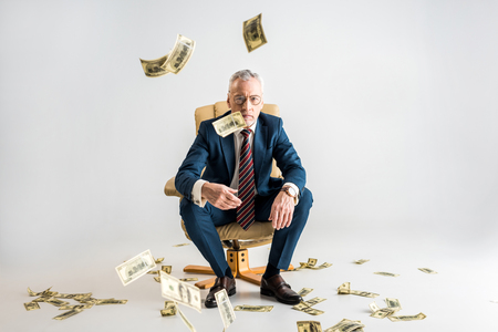 selective focus of mature businessman sitting in armchair and throwing in air dollar banknotes on greyの写真素材