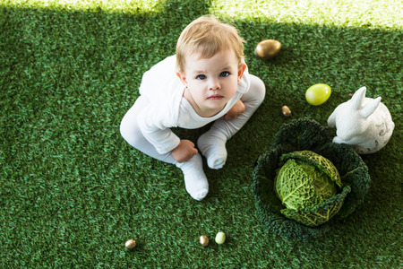 overhead view of cute kid looking at camera while sitting near Easter eggs, decorative rabbit and...