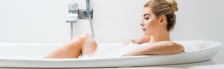 panoramic shot of beautiful and blonde woman taking bath with foam and looking away in bathroom