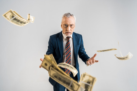 selective focus of mature businessman in glasses and suit throwing in air dollar banknotes isolated on greyの写真素材