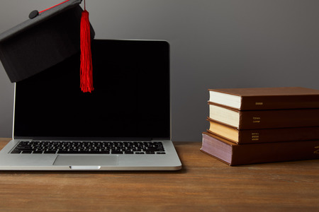 Laptop with blank screen, books and academic cap with red tassel on wooden surface isolated on grey