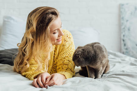 beautiful smiling blonde girl in knitted sweater lying in bed with scottish fold cat at homeの写真素材
