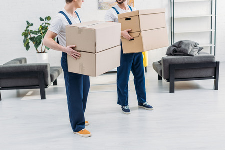 cropped view of two movers in uniform carrying cardboard boxes in apartment