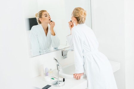 selective focus of beautiful and blonde woman in white bathrobe applying lipstick in bathroomの写真素材