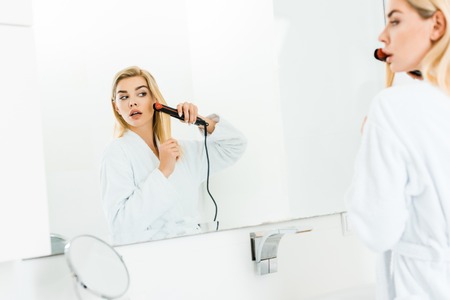 selective focus of beautiful and blonde woman in white bathrobe using flat ironの写真素材
