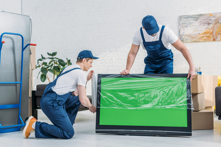 two movers in uniform using roll of stretch film while wrapping tv with green screen in apartmentの写真素材