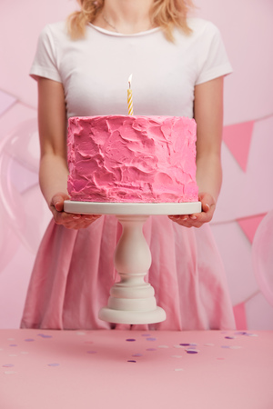 cropped view of woman holding cake stand with sweet pink birthday cake and burning candleの写真素材