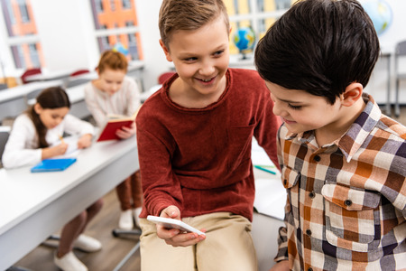 Two smiling schoolboys using smartphone in classroom during brakeの写真素材