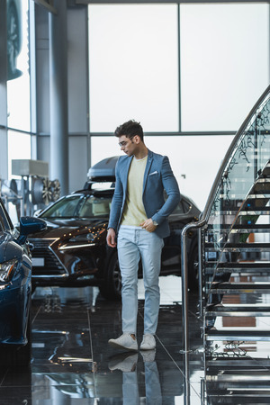 stylish man in glasses standing near automobiles in car showroomの写真素材