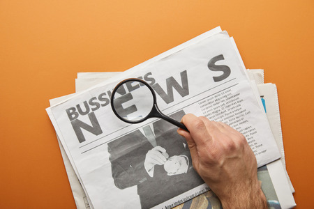 man holding magnifying glass and zooming news lettering while reading  business newspapers on orangeの写真素材
