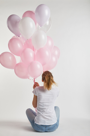 back view of girl in casual clothes sitting and holding pink air balloons on whiteの写真素材