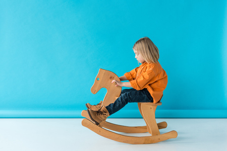 side view of blonde and cute kid riding on rocking horse on blue backgroundの写真素材