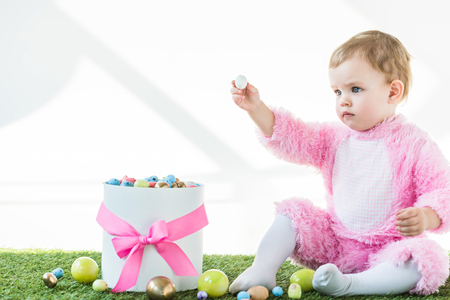 adorable child in pink fluffy costume holding quail egg in outstretched hand while sitting near box with colorful Easter eggs isolated on whiteの写真素材