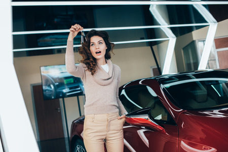 surprised curly woman looking at keys while standing with hand in pocket near red carの写真素材