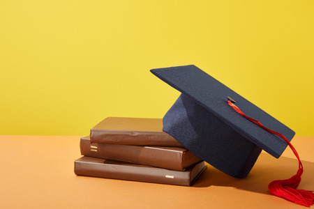 Brown books and academic cap with red tassel on orange surface isolated on yellowの写真素材