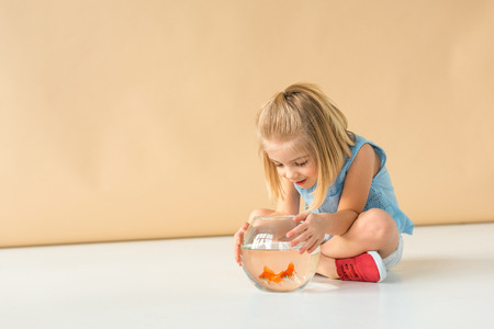 adorable kid sitting with crossed legs and looking at fishbowl on beige backgroundの写真素材