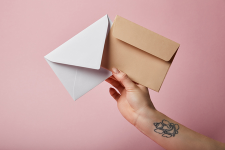 partial view of woman with tattoo holding white and beige envelopes on pink backgroundの写真素材