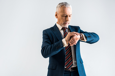 handsome mature businessman in suit standing and looking at watch isolated on greyの写真素材