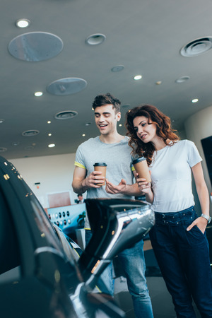 cheerful man holding paper cup near curly woman standing with hand in pocket while looking at automobileの写真素材