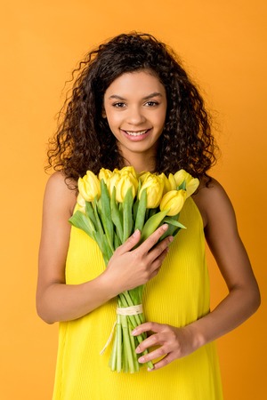 happy curly african american woman holding yellow tulips isolated on orangeの写真素材
