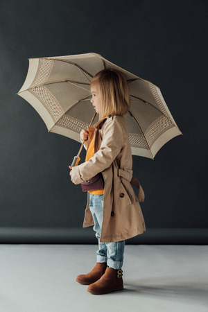 side view of child in trench coat and jeans holding umbrella on black backgroundの写真素材