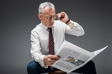 mature businessman touching glasses while reading newspaper on greyの写真素材
