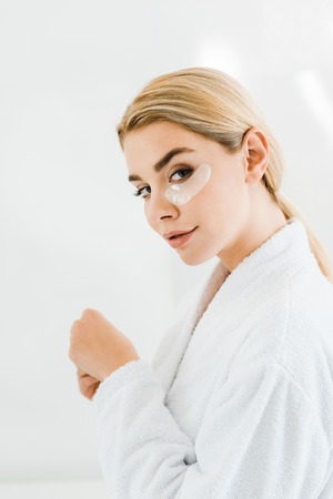 beautiful and blonde woman in white bathrobe with eye patches on face looking at camera in bathroomの写真素材