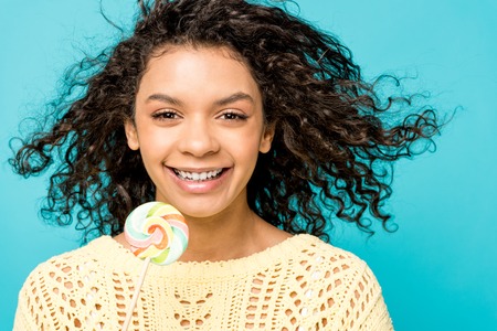 cheerful curly african american girl smiling while holding lollipop isolated on blueの写真素材