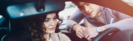 panoramic shot of beautiful curly woman sitting in car and holding steering wheel near cheerful man in glassesの写真素材