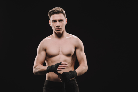 strong shirtless muscular mma fighter fixing bandages and looking at camera isolated on blackの写真素材