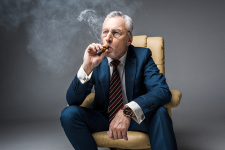 mature man in glasses and suit smoking cigar while sitting in armchair on greyの写真素材