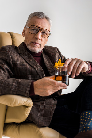 stylish mature man sitting in armchair and holding glass of whiskey isolated on greyの写真素材