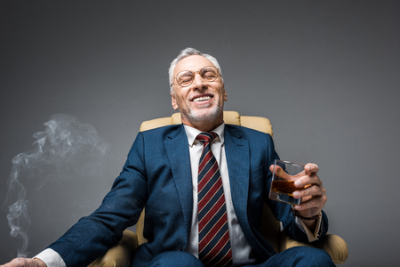 smiling mature businessman in suit holding glass of whiskey while sitting in armchair on greyの写真素材