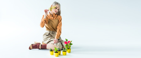 panoramic shot of beautiful stylish girl posing with flower pots and fern leaf on white
