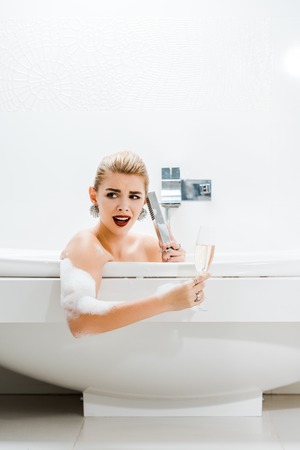 beautiful and blonde woman taking bath, holding champagne glass and talking in bathroom