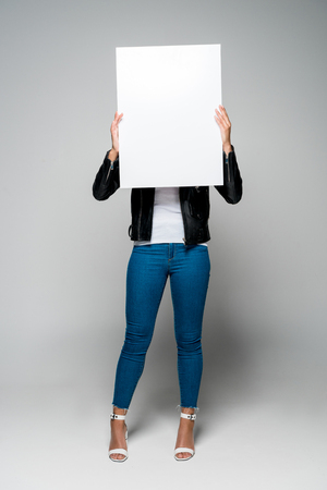 african american young woman covering face with blank placard while standing on greyの写真素材