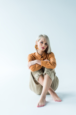 beautiful stylish young woman sitting with crossed legs and looking at camera on whiteの写真素材