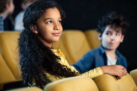 selective focus of two multicultural children sitting in cinema and watching movieの写真素材
