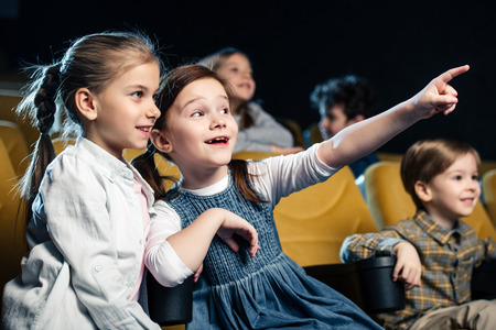 excited kid pointing with finger while watching movie in cinema together with multicultural friendsの写真素材