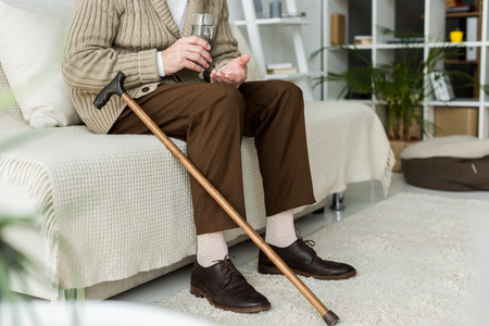 cropped view of retired man holding pill while sitting on sofa near walking caneの写真素材