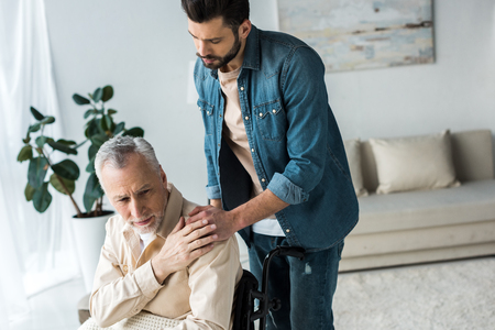 caring handsome son holding hands with disabled senior father in wheelchair at homeの写真素材