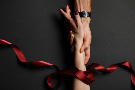 cropped view of man holding female hand in red satin ribbon on black backgroundの写真素材