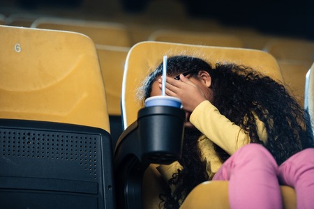 scared african american child holding hand on face while sitting in cinema seat with paper cup in cup holderの写真素材