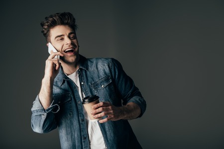 smiling and brunette man in denim shirt holding paper cup and talking on smartphone isolated on blackの写真素材