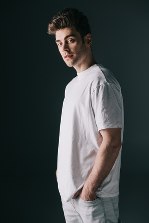 handsome man in white t-shirt and jeans with hands in pockets looking at camera isolated on blackの写真素材